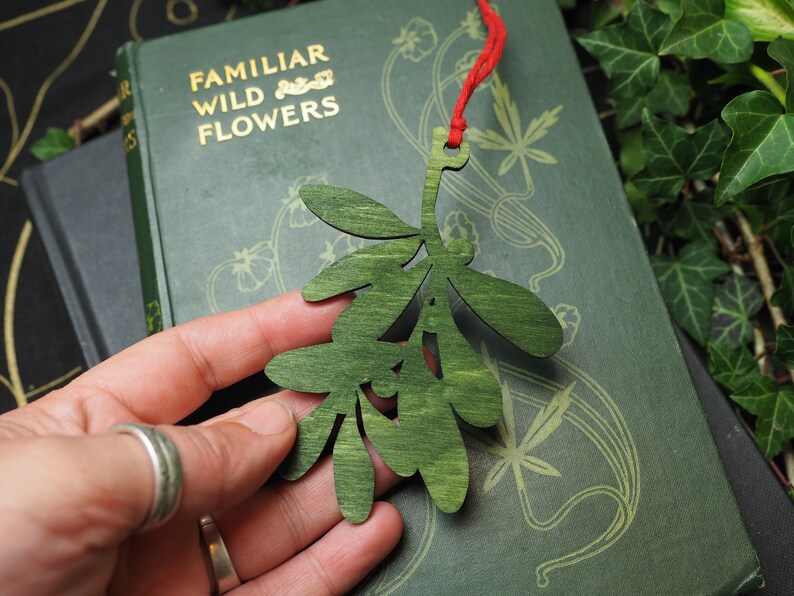 May include: A green wooden mistletoe sprig ornament with a red string attached. The ornament is held in a hand against a green book with the text "Familiar Wild Flowers" on the cover.