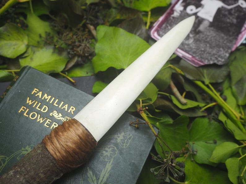 May include: A handcrafted wand with a cream-colored tip, wrapped with brown twine, and a dark wooden handle. The wand rests on a vintage book titled "Familiar Wild Flower" with gold lettering, surrounded by green leaves and a small black and white photograph.