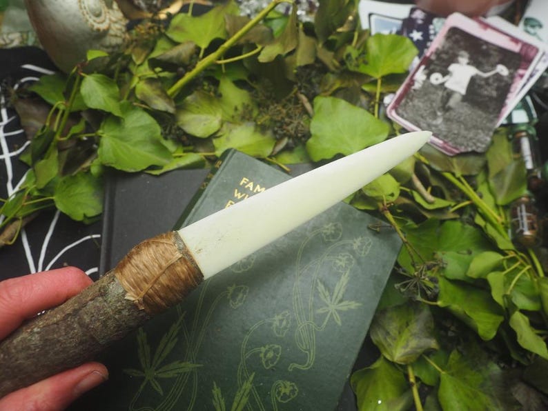 May include: A handcrafted wand with a light-colored, pointed tip and a wooden handle wrapped with natural fiber. The wand rests on a dark green book with botanical illustrations, surrounded by green leaves and other mystical items.