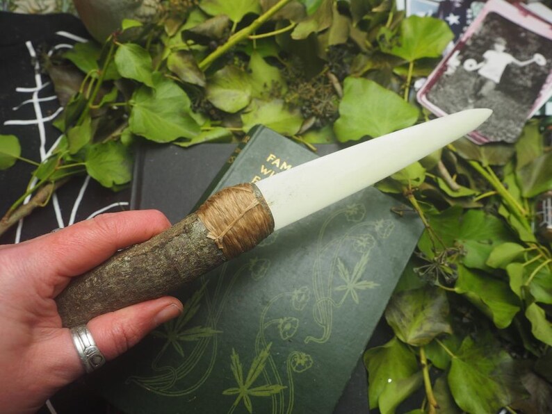 May include: A handcrafted ritual wand with a wooden handle and a creamy white, pointed tip. The handle is wrapped with natural twine. The wand rests on a green book with botanical illustrations, surrounded by green foliage.