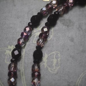 May include: A necklace featuring two strands of faceted beads in alternating shades of deep black and translucent purple. The beads vary in size and shape, creating a textured appearance along the length of the strands, which are arranged in parallel.