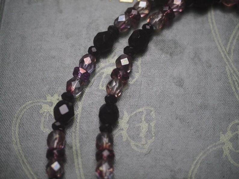 May include: A necklace featuring two strands of faceted beads in alternating shades of deep black and translucent purple. The beads vary in size and shape, creating a textured appearance along the length of the strands, which are arranged in parallel.
