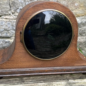 May include: A vintage wooden clock with a dark, circular face and a brass-colored bezel. The clock has a curved top and a rectangular base with decorative trim. The wood has a warm, brown tone, and the clock appears to be in good condition.