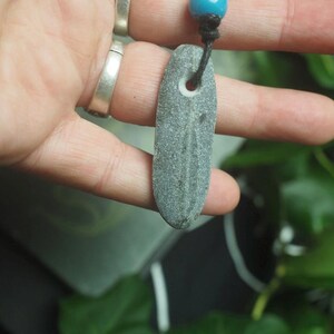 May include: A gray, elongated, flat stone pendant with a natural hole near the top, suspended from a black cord featuring a single, round, bright blue bead. The stone has a smooth, weathered texture and a subtle, darker vertical line running down its center.