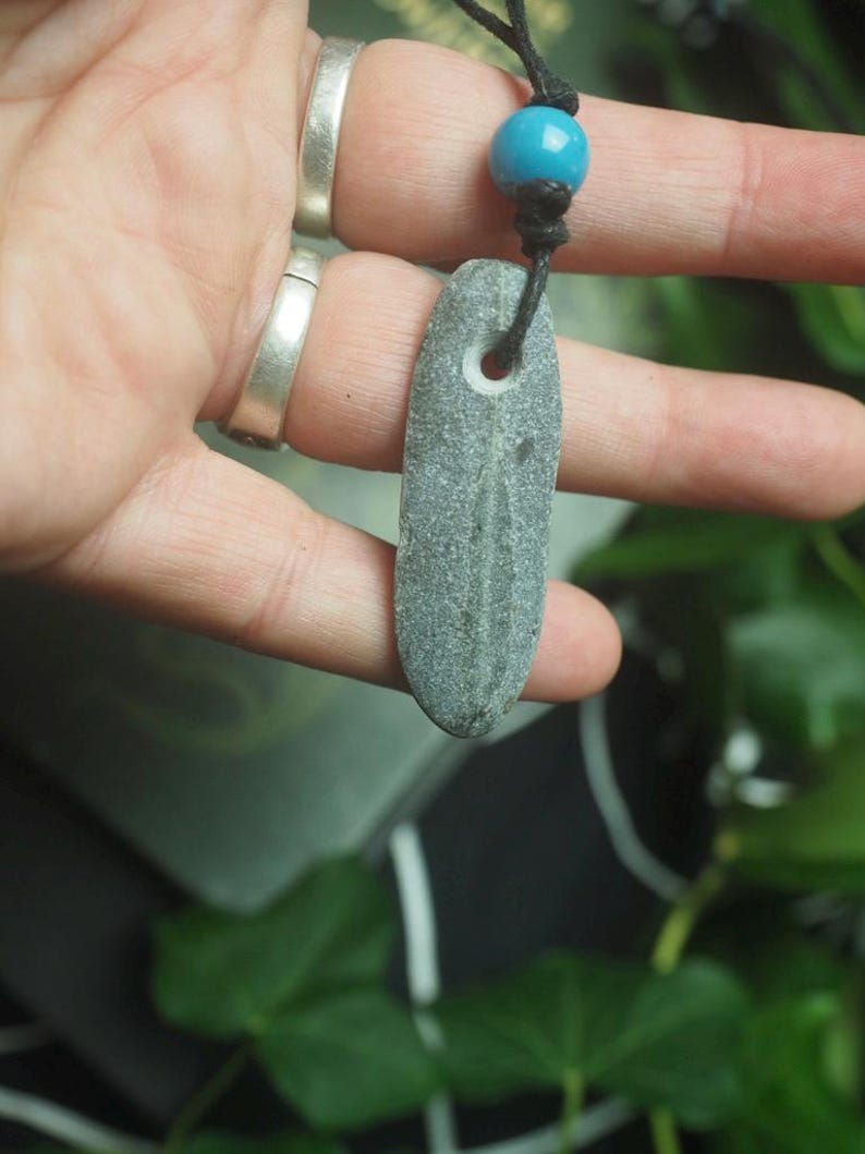 May include: A gray, elongated, flat stone pendant with a natural hole near the top, suspended from a black cord featuring a single, round, bright blue bead. The stone has a smooth, weathered texture and a subtle, darker vertical line running down its center.