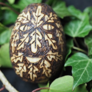 May include: A wooden egg with a carved design of a face made of leaves. The egg is brown and the leaves are carved in a light brown color.