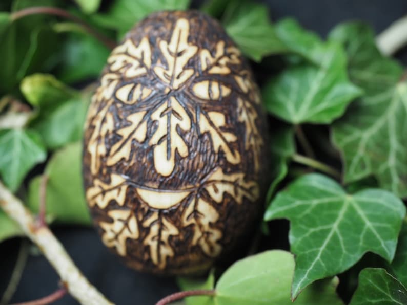 May include: A wooden egg with a carved design of a face made of leaves. The egg is brown and the leaves are carved in a light brown color.