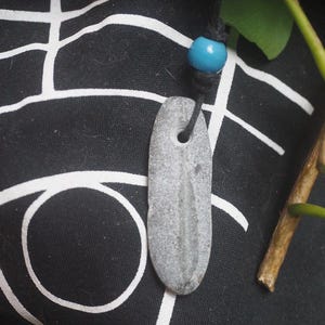 May include: A gray, elongated, smooth stone pendant with a natural hole near the top, suspended from a black cord featuring a single, round, bright turquoise blue bead. The stone has a subtle, vertical, lighter-colored linear marking.