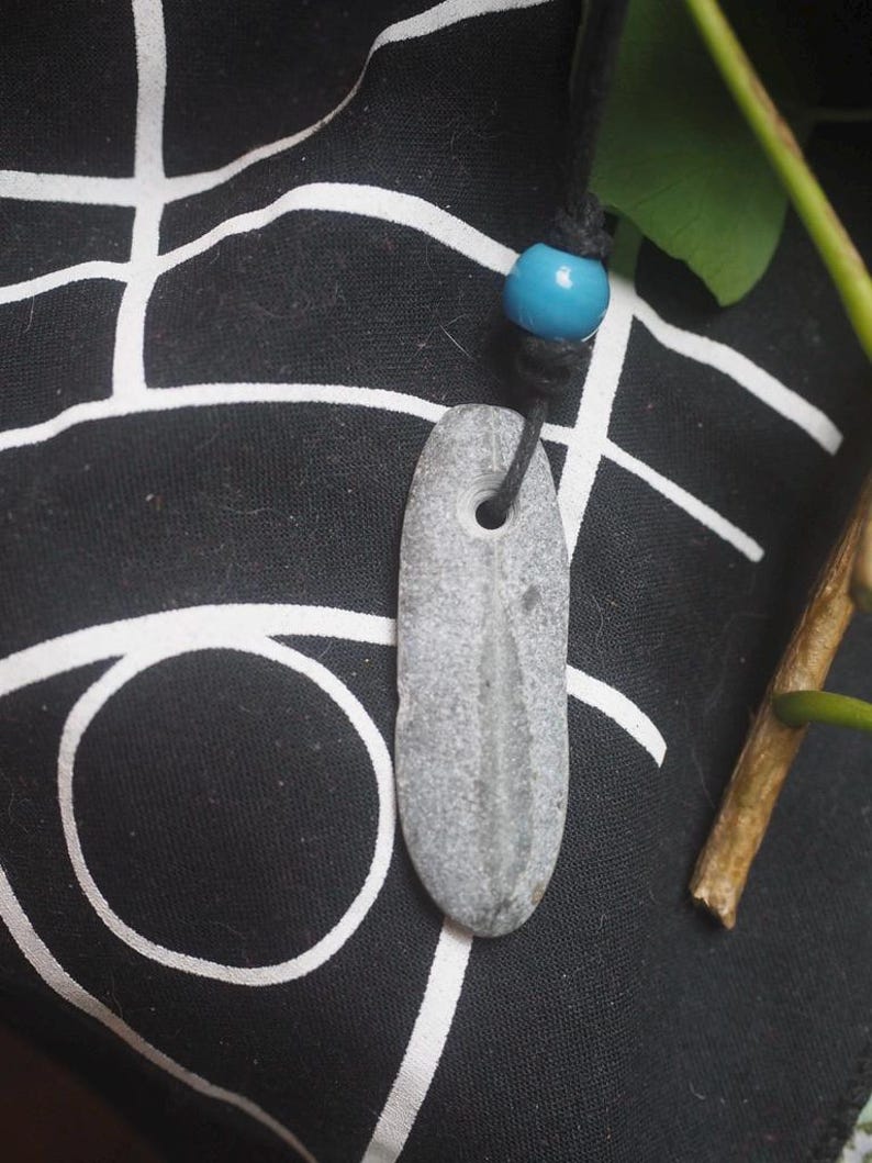 May include: A gray, elongated, smooth stone pendant with a natural hole near the top, suspended from a black cord featuring a single, round, bright turquoise blue bead. The stone has a subtle, vertical, lighter-colored linear marking.