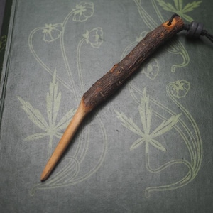 May include: A wooden stick with a dark brown bark-like texture, attached to a black cord with a bead. The stick is lying on a green book with a floral design.