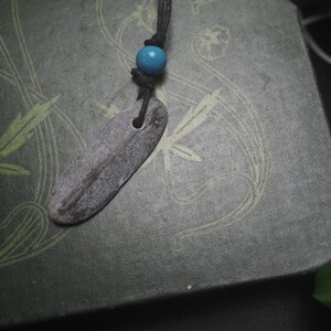 May include: A necklace featuring a flat, elongated, grey stone pendant with a natural texture, suspended from a black cord. A single, smooth, bright turquoise blue bead is threaded onto the cord just above the stone pendant, adding a pop of color.