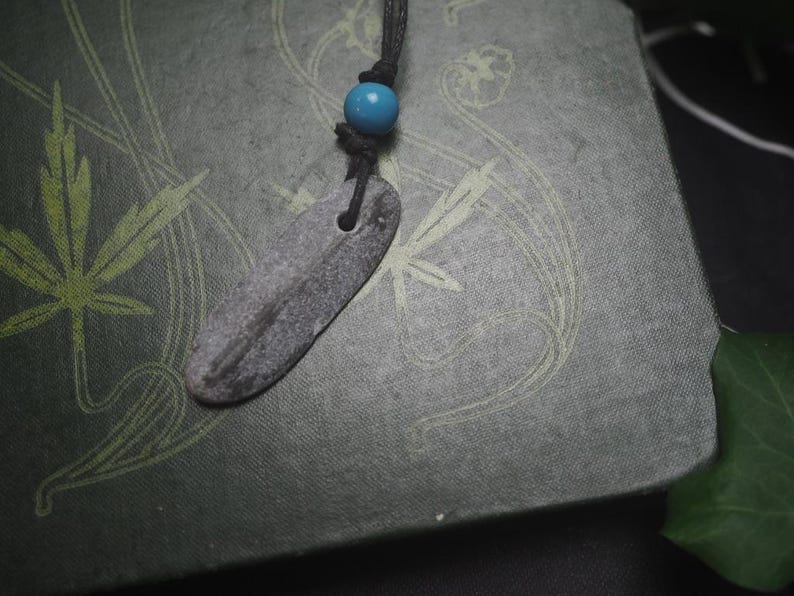 May include: A necklace featuring a flat, elongated, grey stone pendant with a natural texture, suspended from a black cord. A single, smooth, bright turquoise blue bead is threaded onto the cord just above the stone pendant, adding a pop of color.