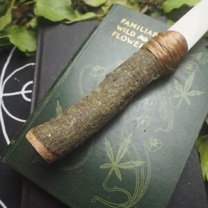 May include: A handcrafted wand with a natural wood handle and a white tip, bound with brown twine. The wand rests on a vintage green book with gold lettering and botanical illustrations. The background includes greenery and a black patterned surface.