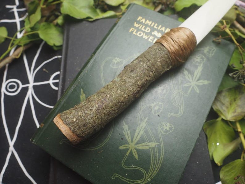 May include: A handcrafted wand with a natural wood handle and a white tip, bound with brown twine. The wand rests on a vintage green book with gold lettering and botanical illustrations. The background includes greenery and a black patterned surface.