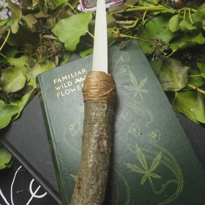 May include: A handcrafted ritual athame with a wooden handle and a cream-colored blade. The handle is wrapped with twine. The athame rests on a vintage book with the title "Familiar Wild Flowers" and is surrounded by greenery.
