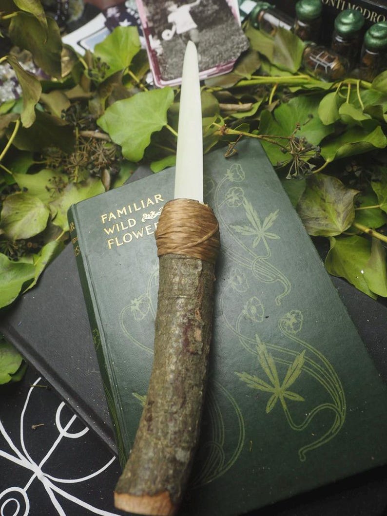 May include: A handcrafted ritual athame with a wooden handle and a cream-colored blade. The handle is wrapped with twine. The athame rests on a vintage book with the title "Familiar Wild Flowers" and is surrounded by greenery.