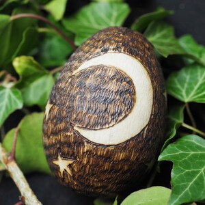 May include: A wooden egg with a crescent moon and star design burned into the surface. The egg is brown with white accents and is surrounded by green leaves.