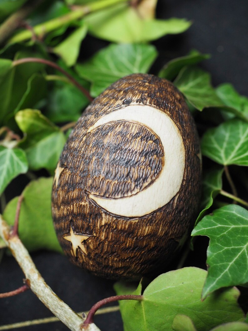 May include: A wooden egg with a crescent moon and star design burned into the surface. The egg is brown with white accents and is surrounded by green leaves.