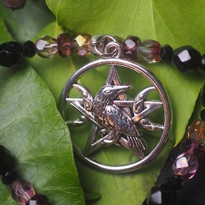 May include: A silver-toned circular pendant featuring a central raven perched on a pentagram, suspended from a beaded necklace. The necklace consists of alternating faceted black beads and translucent purple-tinted beads strung together.