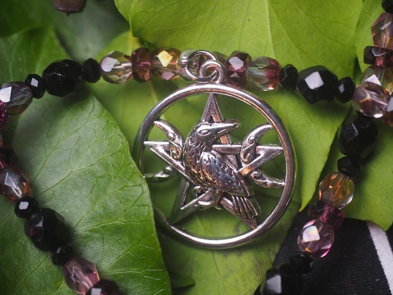 May include: A silver-toned circular pendant featuring a central raven perched on a pentagram, suspended from a beaded necklace. The necklace consists of alternating faceted black beads and translucent purple-tinted beads strung together.