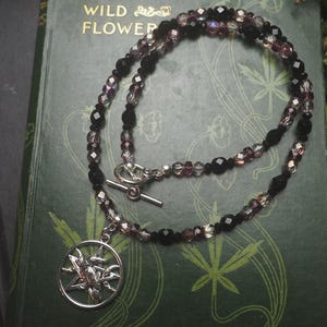 May include: A necklace featuring a circular silver-toned pendant with a raven perched on a pentagram. The chain consists of alternating faceted black and translucent purple beads, secured with a silver-toned toggle clasp. The design is intricate.