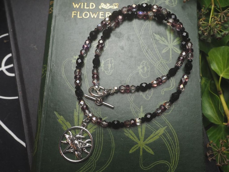 May include: A necklace featuring a circular silver-toned pendant with a raven perched on a pentagram. The chain consists of alternating faceted black and translucent purple beads, secured with a silver-toned toggle clasp. The design is intricate.