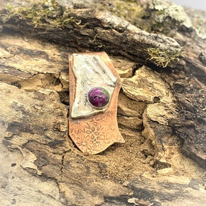 May include: A copper and silver pendant with a textured surface and a round, green and purple gemstone set in the center.