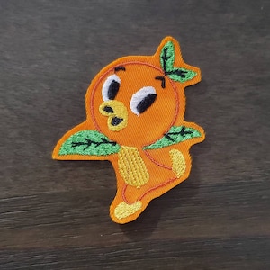Könnte beinhalten: Gestickter Aufnäher mit einer Cartoon-Orange mit grünen Blättern und einem gelben Schnabel. Die Orange hat schwarze Augen und einen weißen Glanz.