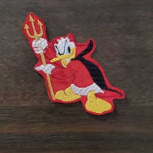 Peut inclure: Patch brodé rouge représentant un canard de dessin animé avec une expression diabolique tenant un trident. Le canard porte une cape rouge avec une doublure noire.