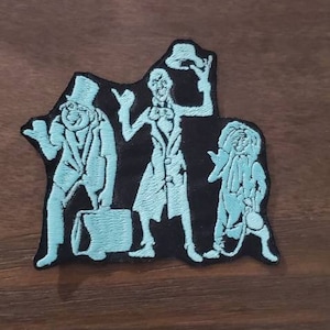 Puede incluir: Un parche bordado con tres figuras fantasmales de la atracción Haunted Mansion en Disneyland. Las figuras son de color azul claro y están delineadas en negro. El parche está sobre una superficie de madera marrón.