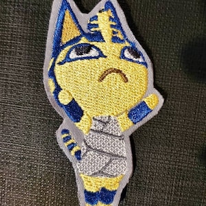 May include: An embroidered patch featuring a cartoon cat with a pharaoh headdress. The cat is yellow with blue accents and is wearing a gray and white striped shirt. The patch is on a dark gray background.