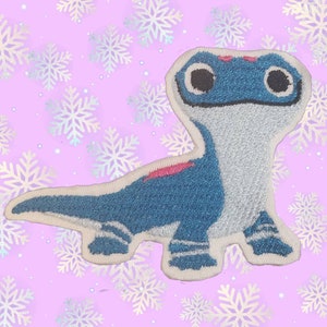 May include: A blue and white embroidered patch of a cartoon lizard with pink accents. The lizard has large eyes and a friendly expression.