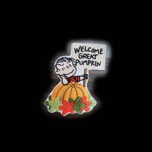 Può includere: Toppa ricamata con un personaggio dei cartoni animati che tiene in mano un cartello con la scritta "WELCOME GREAT PUMPKIN". Il personaggio è in piedi su una grande zucca arancione con foglie verdi e rosse. La toppa ha uno sfondo nero.
