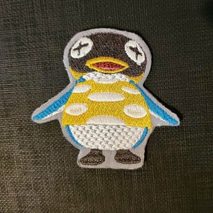May include: Embroidered patch of a penguin wearing a yellow and white polka dot shirt with blue accents. The penguin has a black head and a red beak.