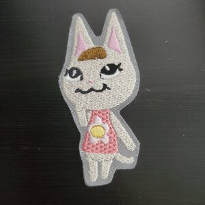 May include: White cat with brown hair, wearing a pink dress with a yellow flower, embroidered patch.