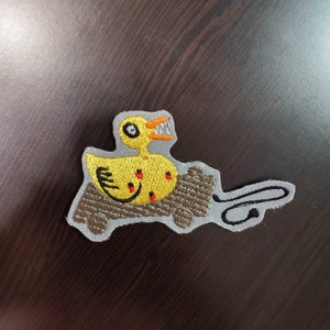 Puede incluir: Un parche bordado que representa un pato amarillo con una sonrisa de dientes, sentado en un tronco de madera gris bordado. El pato tiene puntos rojos en su cuerpo.