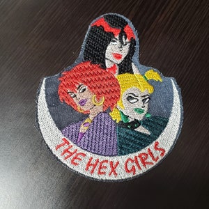 Op de afbeelding: Gestikt patch met drie cartoonvrouwen met kleurrijk haar en kleding. Het patch is rond en heeft de tekst "The Hex Girls" in witte borduurwerk.
