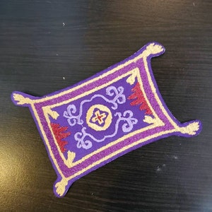 Puede incluir: Un parche de alfombra mágica bordado en color morado y dorado con un diseño rojo y dorado en el centro.