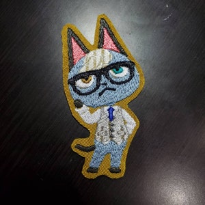 May include: An embroidered patch of a blue cat wearing glasses and a white shirt with a blue tie. The cat has a yellow and white striped headband and a gold outline.