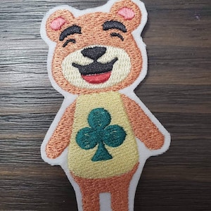 May include: Embroidered patch of a brown bear with a yellow shirt and a green clover. The bear is smiling with its eyes closed.