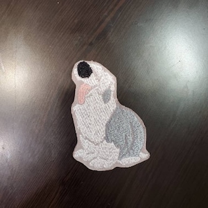 May include: Embroidered iron-on patch of a white and gray dog sitting with its head tilted. The dog has a black nose and pink tongue.