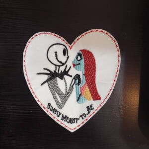 Puede incluir: Parche bordado en forma de corazón con Jack Skellington y Sally de Pesadilla Antes de Navidad. El parche es blanco con costuras rojas alrededor de los bordes y el texto "Simply Meant To Be" en la parte inferior.