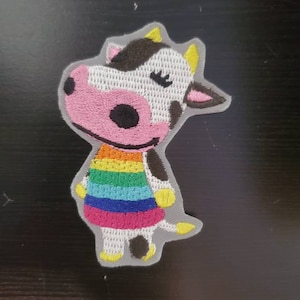 May include: Embroidered patch of a white cow with black spots wearing a rainbow striped dress. The cow has a pink nose and black eyes with eyelashes.