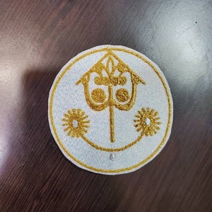 May include: A white embroidered patch with a gold design. The design features a stylized castle with a tower and a three-pointed crown on top. There are two stylized flowers on either side of the castle.
