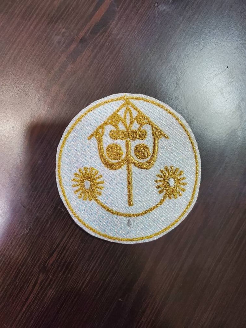 Puede incluir: Un parche bordado blanco con un dise&ntilde;o dorado. El dise&ntilde;o presenta un castillo estilizado con una torre y una corona de tres puntas en la parte superior. Hay dos flores estilizadas a cada lado del castillo.