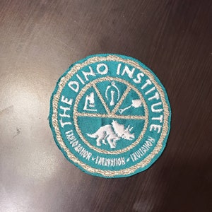 Puede incluir: Un parche bordado azul turquesa con el texto "The Dino Institute" en blanco. El parche presenta un dinosaurio blanco y un símbolo con una pala, un hueso y una lupa.
