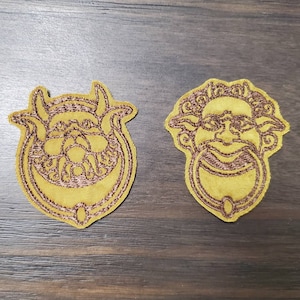 Puede incluir: Dos parches bordados con un diseño dorado de la cara de una criatura mítica. Los parches están sobre una superficie de madera marrón.