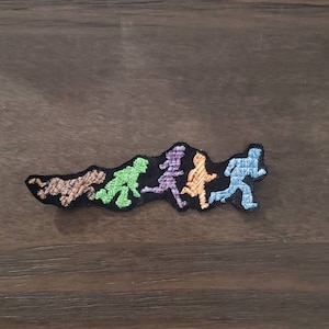 Może przedstawiać: Naszywka haftowana przedstawiająca linię postaci w różnych kolorach, w tym pomarańczowym, zielonym, fioletowym i niebieskim, biegnących w linii. Postacie są stylizowane i reprezentują postęp ewolucji człowieka.