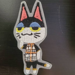 May include: Embroidered patch of a black and white cat with yellow eyes, wearing a plaid shirt. The cat has a gray background and is standing with its tail curled behind it.