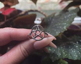 Puede incluir: Un anillo de plata con un diseño de pentagrama. El anillo está en un dedo y el fondo está desenfocado.